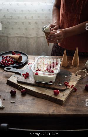 Abgeschnittene, nicht erkennbare Frau, die einen Kegel mit hausgemachtem Eis hält, mit Johannisbeeren und Stachelbeeren auf einem rustikalen Tisch. Stockfoto