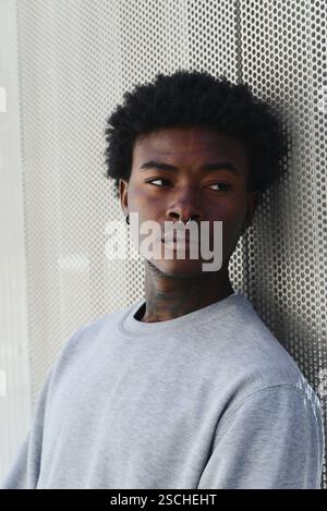 Porträt eines jungen schwarzen Mannes mit Gesichtspiercings und Tattoos, der einen legeren grauen Pullover trägt und sich an eine strukturierte Metallwand lehnt. Er ist loo Stockfoto