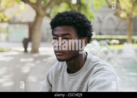 Porträt eines jungen schwarzen Mannes, der draußen in einem Park sitzt und mit einem nachdenklichen Ausdruck wegblickt. Er trägt einen lässigen grauen Pullover und trägt eine Nase Stockfoto