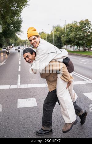 Ein fröhliches kolumbianisches Schwulenpaar teilt einen verspielten Moment mit einer Huckepack-Fahrt auf einer Straße der Stadt. Üppige Bäume säumen den Straßenrand und schaffen eine lebhafte Stadt Stockfoto