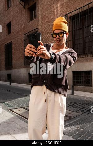 Ein modischer lateinmann in gelber Beanie, Sonnenbrille und lässiger Kleidung macht ein Selfie mit einem Smartphone in einer urbanen Umgebung und zeigt einen modernen an Stockfoto