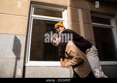 Ein glückliches schwules Paar in stilvoller Kleidung feiert die Liebe an einem sonnigen Tag. Die urbane Kulisse unterstreicht ihre fröhliche Verbindung und verspielte Energie, Capturi Stockfoto