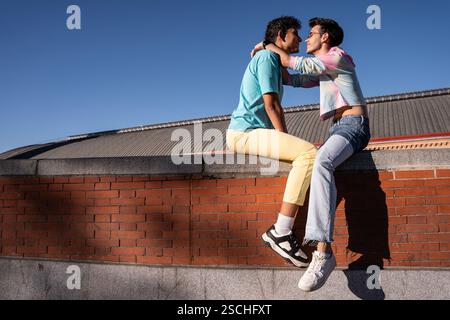 Ein freudiger Moment, den ein schwules Paar teilt, das auf einer Ziegelmauer unter einem klaren blauen Himmel sitzt. Ihre lebhafte Interaktion und farbenfrohe Kleidung spiegeln das Glück wider Stockfoto