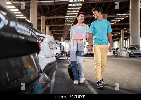 Ein schwules Paar geht Hand in Hand durch einen sonnigen Parkplatz voller Autos. Beide lächeln und genießen einen Moment der Zweisamkeit und symbolisieren lov Stockfoto