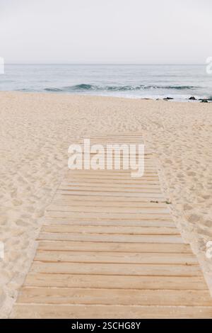 Eine hölzerne Promenade erstreckt sich über Sanddünen, die zu einem Strand führt, an dem sanfte Wellen auf das Ufer treffen und eine friedliche Küstenlandschaft schaffen, die perfekt für Ruhe ist Stockfoto