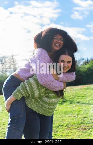 Zwei Freunde teilen sich eine fröhliche Huckepack-Fahrt in einem üppigen, sonnigen Park. Das Lachen und die warmen Pullover sorgen für eine gemütliche, lebendige Atmosphäre, in der die Pflege zum Ausdruck kommt Stockfoto