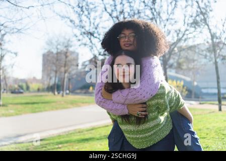 Zwei Freunde teilen einen fröhlichen Moment mit einem Huckepack-Ritt in einem sonnigen Park. Sie tragen farbenfrohe Pullover, die Glück und Freundschaft gegen ein G symbolisieren Stockfoto
