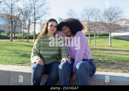 Zwei Freunde in farbenfrohen Pullis sitzen auf einer Parkbank und genießen einen sonnigen Tag. Die pulsierenden grünen und violetten Pullover stehen im Kontrast zum ruhigen Outdoor-bac Stockfoto