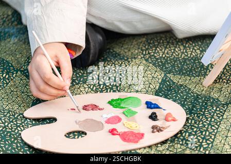 Eine Nahaufnahme einer jungen schwarzen Frau, die eine Holzpalette mit bunten Farbtuben und Pinseln auf einer gemusterten Matte vorbereitet, die Kreativität und artis zum Ausdruck bringt Stockfoto