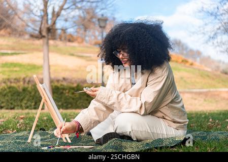 Eine junge schwarze Frau mit lockigen Haaren konzentriert sich auf das Malen in einem ruhigen Park, umgeben von lebhaftem grünem Gras und Bäumen, die Kreativität und relaxa einfangen Stockfoto