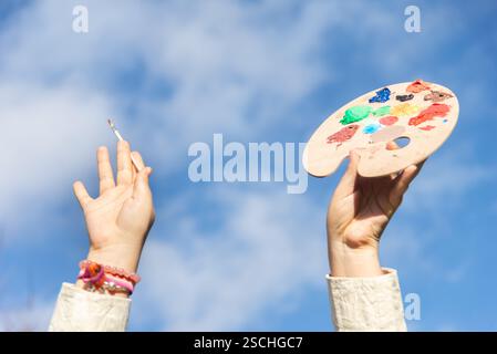 Eine Hand hält eine farbenfrohe Künstlerpalette mit verschiedenen Farben, während eine andere Hand mit einem Pinsel vor einem leuchtend blauen Himmel steht Stockfoto
