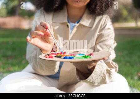 Ein junger schwarzer Künstler hält einen Pinsel und eine Palette voller lebendiger Farben, die sich mit Plein Air-Malerei beschäftigen. Die Szene fängt Kreativität und AR ein Stockfoto