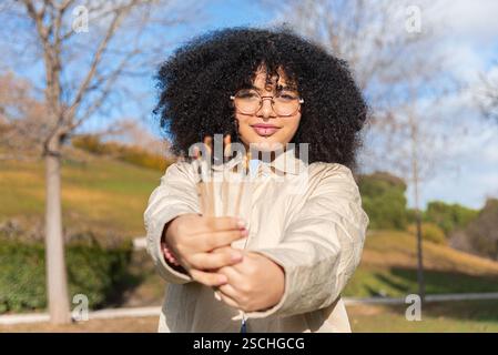 Eine fröhliche schwarze Frau mit Brille und lockigem Haar hält Pinsel in einem sonnigen Park. Der Hintergrund ist mit Bäumen und Grünpflanzen geschmückt, die Creativi hervorheben Stockfoto