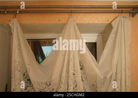 Spitzenvorhänge sind künstlerisch über einem Fenster in einem gemütlichen Wohnzimmer drapiert. Die komplizierten Muster des Stoffes schaffen ein weiches, einladendes Ambiente Stockfoto