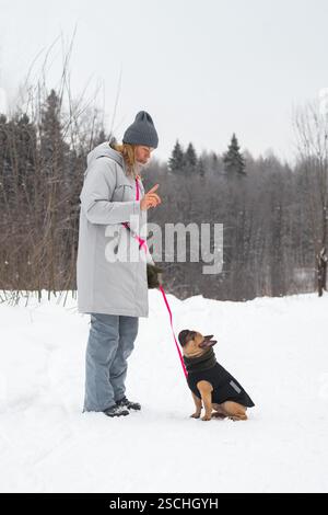 Eine blonde Frau im Wintermantel mit einem Hund an der Leine in einer verschneiten Umgebung. Der Hund sitzt aufmerksam. Schneebedeckte Bäume und ein bewölkter Himmel schaffen eine Stockfoto