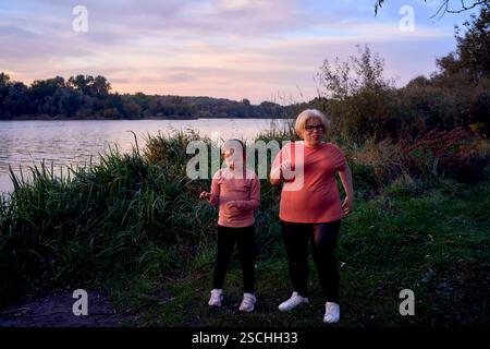 Ein freudiger Moment, in dem Mutter und Tochter in der Abenddämmerung am Fluss tanzen. Das ruhige Wasser und das üppige Grün ergänzen ihren fröhlichen Ausdruck und die Matte Stockfoto