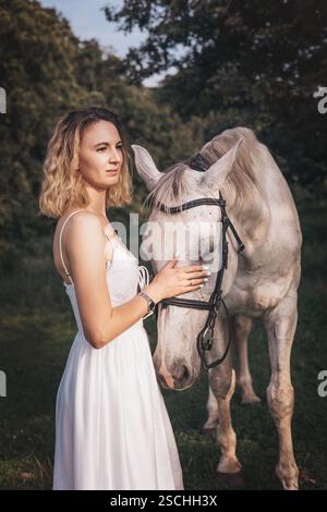 Eine junge Frau in einem fließenden weißen Kleid steht auf einem grasbewachsenen Gebiet und streichelt sanft ein weißes Pferd. Die Szene ist ruhig, eine besondere Bindung zwischen uns Stockfoto
