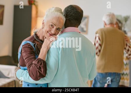 Porträt einer eleganten Seniorin, die in die Kamera schaut, während ihr nicht erkennbarer Partner sie während des Tanzes umarmt Stockfoto