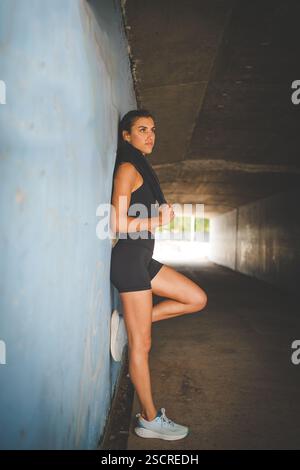 Junge Sportlerin in schwarzer Sportbekleidung, die im Freien verschiedene Fitnessposen vor einem modernen industriellen Hintergrund ausübt. Stockfoto