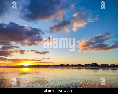 Leuchtende Farben erfüllen den Himmel, während die Sonne hinter einer Küstenstadt untergeht und sich auf das ruhige Wasser reflektiert. Stockfoto