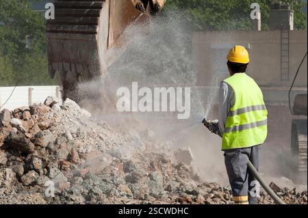 Ein CAT-Bagger sammelt Stahlbetonrückstände. Ein Arbeiter in Sicherheitsausrüstung bewässert ihn mit einem Feuerlöschschlauch, um Staub zu reduzieren. Stockfoto