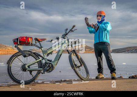 Ein enior-Radfahrer zieht warme Handschuhe an, bevor er mit einem E-Bike am Ufer des Frozen Horsetooth Reservoir in den Ausläufern von Colorado fährt. Stockfoto