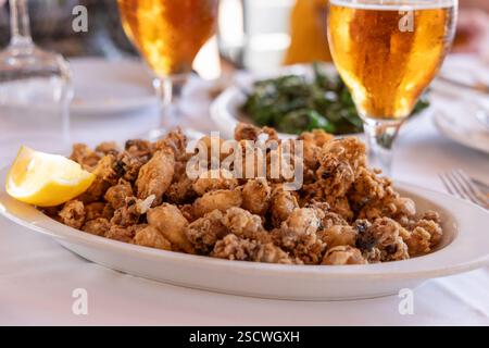 Goldener, knuspriger mediterraner gebratener Calamari (Chipirones), serviert mit einer Zitronenscheibe, Getränken und Grünzeug in einer lockeren Essensumgebung. Stockfoto