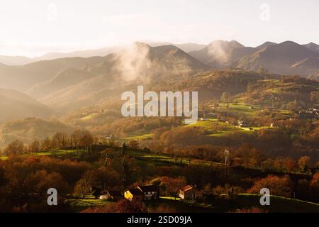 Nebliger Sonnenaufgang über einem Tal mit herbstlichen Farben, Häuser verstreut auf den Hügeln und Berge im Hintergrund. Stockfoto