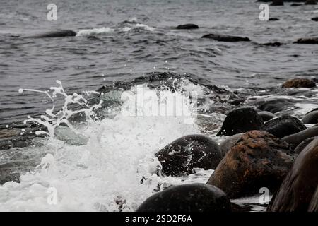 Eine dynamische Nahaufnahme von Meereswellen, die auf glatte, dunkle Kieselsteine an einer zerklüfteten Küste entlang der norwegischen Südküste prallen. Das Wasser spritzt. Stockfoto