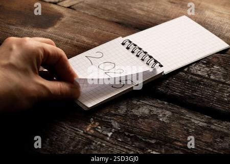 Eine Frauenhand dreht die Seiten eines Notizbuchs, auf dem die Jahre von 2024 bis zum neuen 2025 am Tisch geschrieben sind, der Beginn des neuen 2025, neu Stockfoto