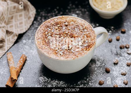 Tasse Cappuccino mit Zimt, Nüssen und Puderzucker auf dunkler rustikaler Oberfläche mit Kaffeebohnen Stockfoto