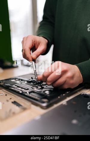 Techniker, männlich, repariert Laptop-Mainboard mit Schraubendreher, umgeben von verschiedenen Komponenten und Werkzeugen auf Werkbank, Nahaufnahme. Computerkonzept Stockfoto