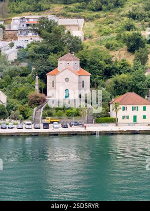 Bucht von Kotor, Montenegro - 2. Juli 2024: Muo Village. Nahaufnahme der historischen Kirche Marias Hilfe der Christen mit Autos, die entlang des Wassers geparkt sind Stockfoto