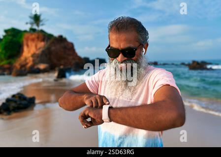 Grauer, bärtiger Seniorenmann mit Sonnenbrille benutzt am Strand eine intelligente Uhr. Reifer Mann mit Ohrstöpseln überprüft Fitness-Tracker auf Gesundheit. Drahtlose Technologie wird verwendet von Stockfoto
