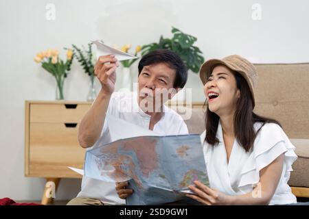 Glückliches asiatisches Paar plant ihre Reise, erkundet eine Karte und teilt spielerische Momente mit einem Papierflugzeug. Stockfoto