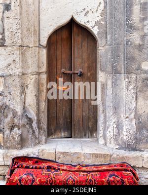 Ein kunstvoller Eingang in einer strukturierten Steinmauer mit komplizierten Schnitzereien, ein lebendiger roter Teppich auf Kopfsteinpflaster, unterstreichen den Charme des alten Baku, Aserbaidschan, während des ersten nationalen Teppichfestes Stockfoto