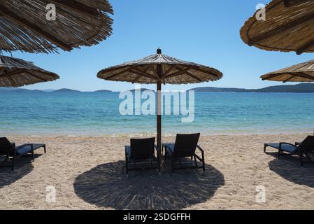 Sonniger Strand mit Liegestühlen unter Strohschirmen. Türkisfarbenes Meer mit Inseln im Hintergrund, entspannte Urlaubsatmosphäre, Amadria Park, White Beach Stockfoto