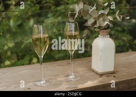 Zwei Sektflöten stehen bereit für eine Feier, begleitet von einem einfachen Eukalyptus-Arrangement in einer dekorierten Vase auf einem Holztisch in einer Natur Stockfoto
