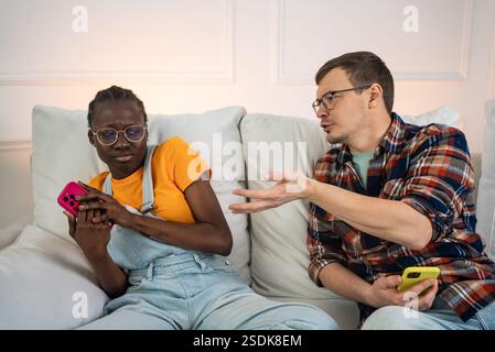 Junge interrassische Paare sitzen auf einem Sofa und haben eine Meinungsverschiedenheit, während beide Smartphones halten. Sie heben Kommunikationsprobleme in modernen Beziehungen hervor Stockfoto