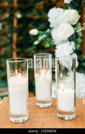 Drei Glaskerzenständer mit weißen Wachskerzen, die sanft brennen, gegen Blumendekorationen und einen rustikalen Holzrahmen bei einer Veranstaltung im Freien. Stockfoto