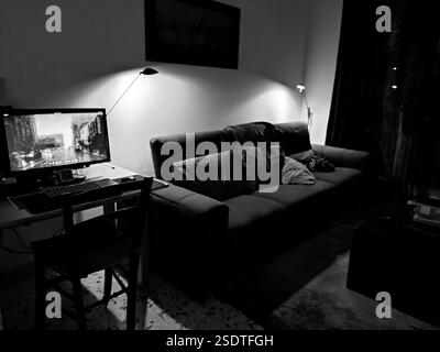 Ein schwach beleuchtetes Zimmer. In der linken Ecke leuchtet ein Computerbildschirm. Eine große Couch mit einer Decke über den Kissen. Stockfoto
