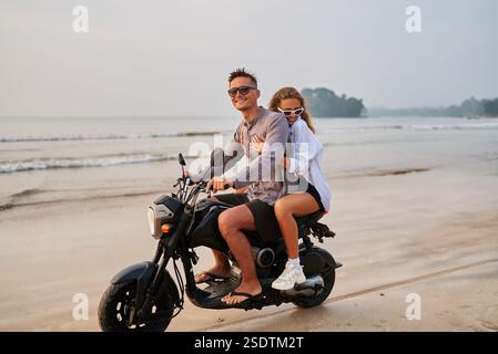 Ein paar Fahrten mit dem Motorrad am Strand entlang. Mann fährt Roller, Frau sitzt dahinter und genießt den sonnigen Blick aufs Meer. Reise-Abenteuer, Erkundung der Küste, Sand Stockfoto
