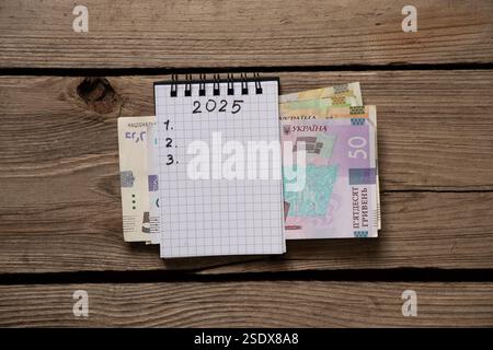 2025 und 123 sind in einem Notizbuch geschrieben und die ukrainische Griwna, die auf einem Holztisch liegt, schreibt Pläne für das neue Jahr auf Stockfoto