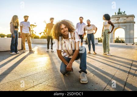 Mann mit lockigen Haaren und lässigen Kleidern, die in die Kamera schaut und neben multiethnischen Freunden in der Stadt während Sonnenuntergang lächelt Stockfoto