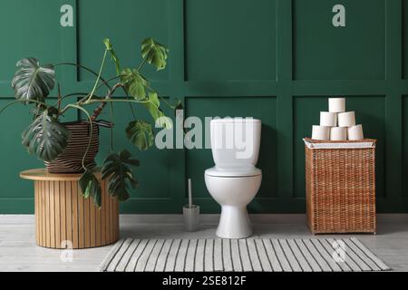 Innere der Toilette mit Toilettenschüssel, Korb mit Papier und Zimmerpflanze in der Nähe der grünen Wand Stockfoto