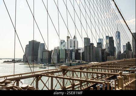 Die Skyline von Manhattan erhebt sich über dem East River, von der Brooklyn Bridge aus gesehen. Helles Sonnenlicht wirft Schatten auf die Struktur der Brücke, während Boote fahren Stockfoto