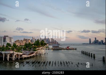 Die Skyline der Stadt erstreckt sich entlang des Ufers, während die Sonne hinter Gebäuden untergeht. Wolken schweben über dem Wasser und werfen sanfte Reflexe auf das Wasser. Stockfoto