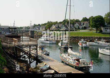 Ein friedlicher Hafen bietet mehrere Segelboote und Fischereifahrzeuge, die in der Nähe eines hölzernen Gehwegs angedockt sind. Charmante Häuser säumen die Uferpromenade, wenn die Sonne scheint Stockfoto