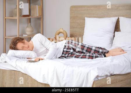 Hübscher junger müder Mann im Pyjama mit Kissen und Buch, der zu Hause auf dem Bett schläft Stockfoto
