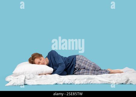 Schöner junger glücklicher Mann im Pyjama mit Kissen und Decke, der auf blauem Hintergrund schläft Stockfoto